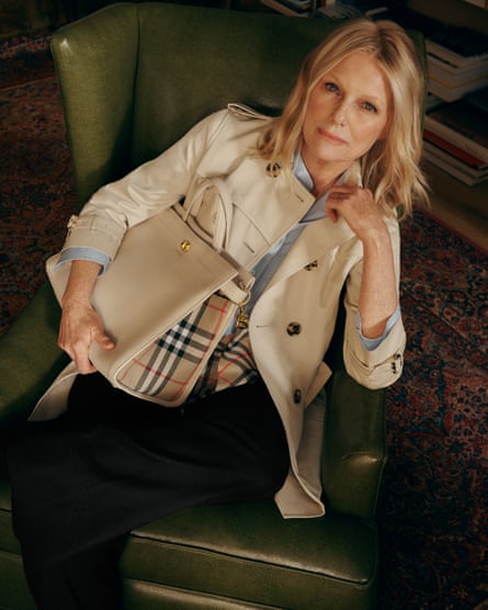 A model wears a short Burberry trench coat while clutching the brand’s Cotswold bag while sitting on green leather armchair