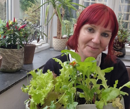 Kim Stoddart holds a long tub of salad leaves as she stands by a windowsill; she has bright red hair and there is a chilli plant behind her.