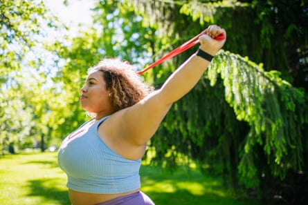 Young woman in purple workout attire stands in park working out with a resistance band