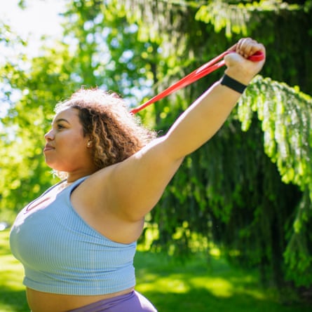A profile view of person with brown skin and curly hair facing to the left in a park holding a red resistance band up in the air and across their back They are wearing a pale blue sleeveless exercise top and are smiling.