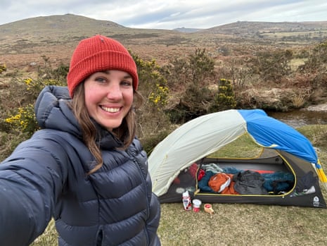 Sian Lewis wearing a black puffer jacket and red beanie takes a selfie with her tent and rolling hills in the background on a wild camping trip
