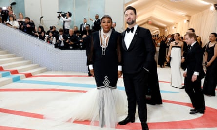 Serena Williams in a slim black dress, with long sleeves and cuffs, that flares out into a white wedding-dress-style train at the bottom. Next to her is Alexis Ohanian in full black tie with a cape