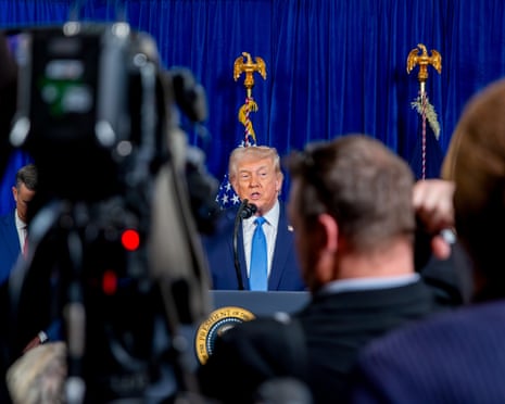 Man in suit at table with blue curtain background, in front of mob of TV crews.