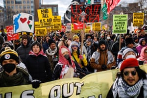 people hold anti-Trump and pro-Palestine signs at a protest, with messages such as 'End all US aid to Israel!' and 'Shut down the anti-migrant Laken Riley Act'
