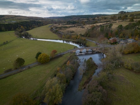 View over landscape showing a bridge over a river, agricultural fields, trees and high moorlands and hills in the background.