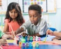 A young boy reaches over a table of paint pots while other children paint nearby