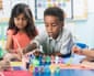 A young boy reaches over a table of paint pots while other children paint nearby