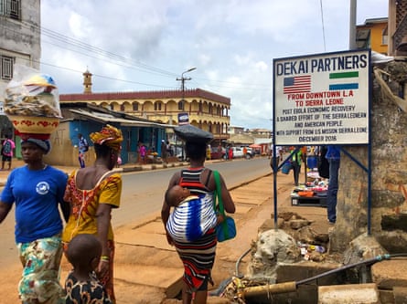 Women walk down a main road in Freetown, Sierra Leone, past a sign saying Dekai Partners. Two are balancing bowls on their heads