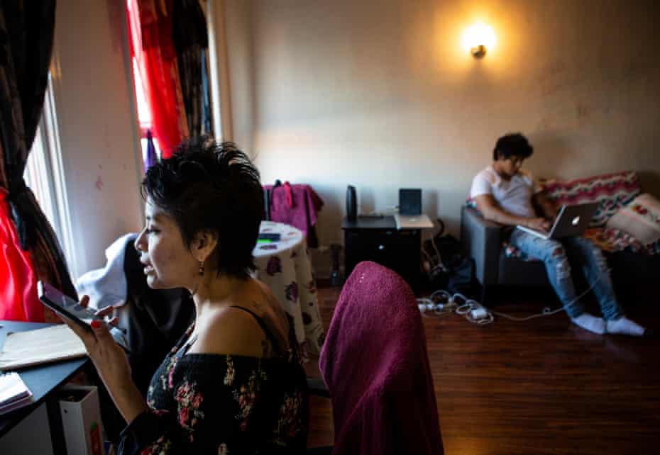A woman with short black hair speaks into a phone, in the background a man uses a laptop
