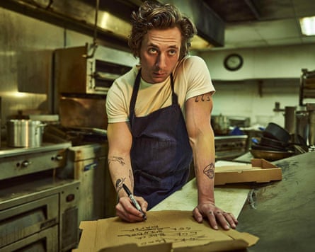 Jeremy Allen White in a white T-shirt and blue apron leans on a counter in a kitchen in a scene from The Bear.