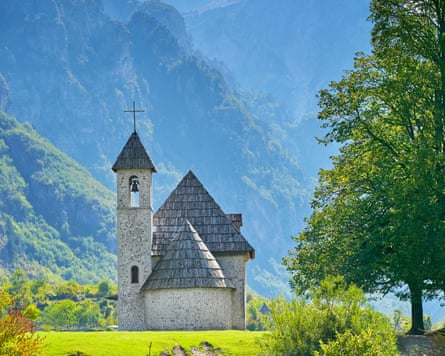 A small church on a sunny day with wooded mountains behind.