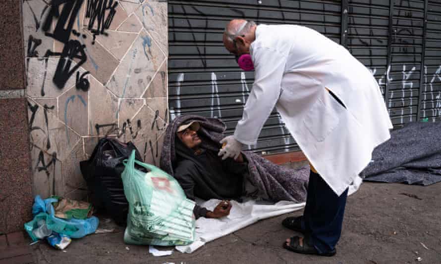 Lancellotti, in his white coat, leans over a homeless man lying in a sleeping bag on the pavement