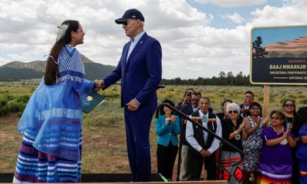 Youth leader Maya Tilousi, wearing blue traditional garb, shakes hands with Joe Biden, who wears a baseball cap with his suit, in front of a small group of people, against a backdrop of green desert brush, a brown peak and white clouds and blue sky.