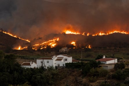 Smoke blots out the sky as flames run the full length of a hilly landscape, with white-walled Mediterranean homes dangerously close by