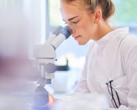 a female research scientist is analysing a sample on her microscope in a microbiology lab. the lab is brightly lit with natural light.
