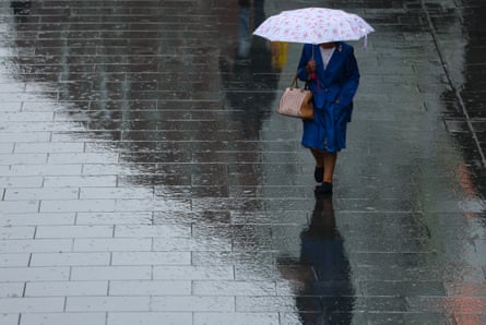 A woman wearing a raincoat and holding an umbrella walks along rain-soaked pavement in Liverpool.
