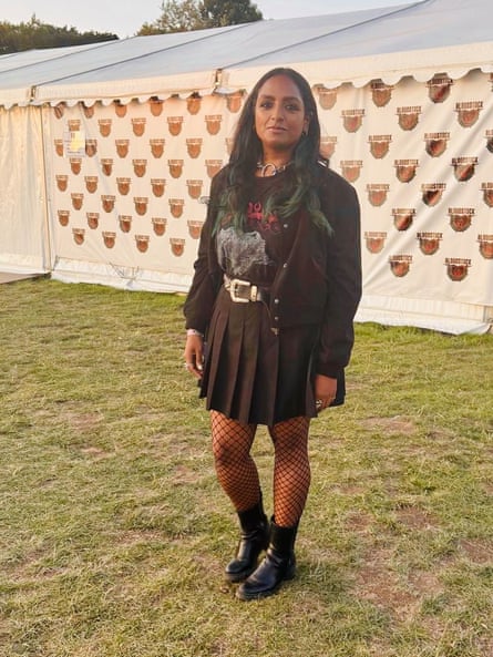 A full length picture of a young woman wearing a short pleated black shirt, fishnet tights, jacket and motif T-shirt and long dark hair and ankle boots - standing on grass in front of a white festival tent
