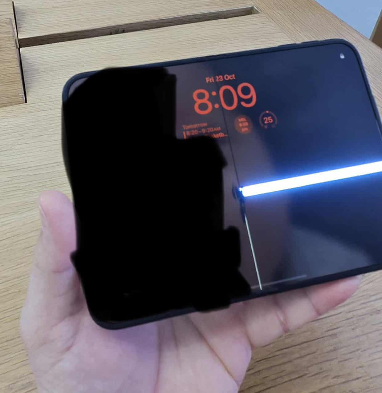 A hand holds a folding iPhone with a visible crack line along its vertical hinge and a white strip of horizontal light across the right-hand side of the screen
