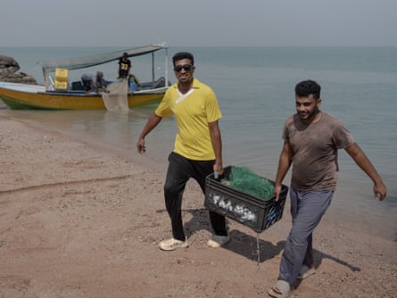 Two fishers carry a box at shoreline with small boat in background
