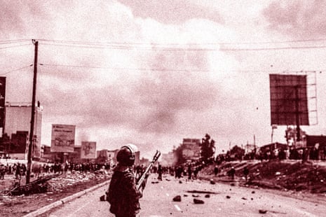 Monochrome photo of a policeman in riot gear holding a teargas grenade launcher on a main road filled with rubble as crowds group in the distance