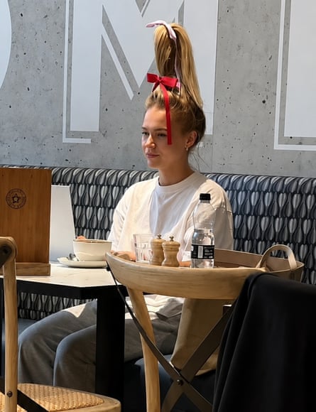 A blond woman in a white t-shirt with an extremely tall ponytail on her head sits in a coffee shop