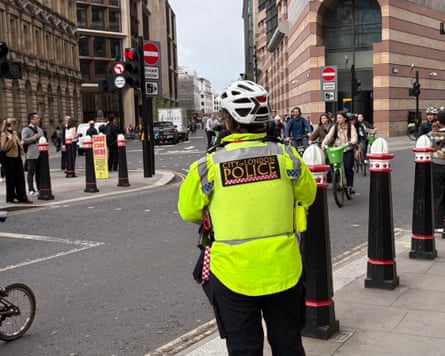 A City of London police cyclist officer stands at a street junction near cyclists and bollards