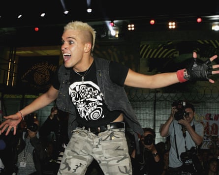 A man with short bleached blond hair stands with his arms outstretched, wearing a black T-shirt, grey jeans and black fingerless gloves.