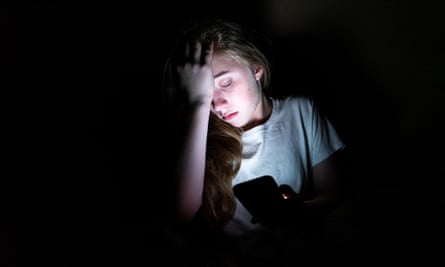 Garota chateada sentada no escuro enquanto usa seu smartphone. A luz da tela ilumina seu rosto.