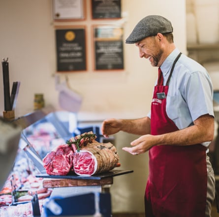 A butcher wearing a flat cap and apron with a joint of meat on the counter