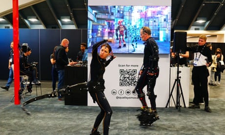 A woman and a man in black bodysuits with small - rivets? - attached to them either stand on machinery or have it attached to their bodies, on the carpeted floor of a high-ceilinged conference hall beside exhibitor booths.