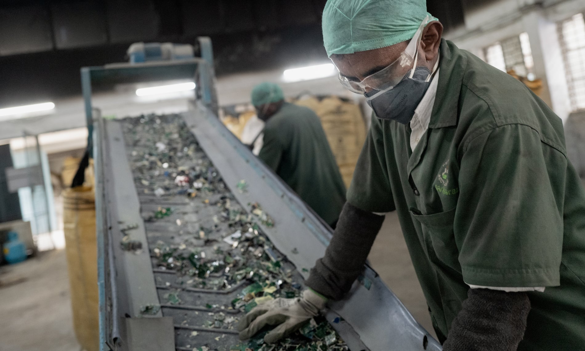 Trabajador con uniforme verde, guantes, mascarilla y gorro de protección, opera una cinta transportadora que lleva desechos electrónicos en una planta de reciclaje. En el fondo, otro trabajador realiza labores similares en un ambiente industrial iluminado.