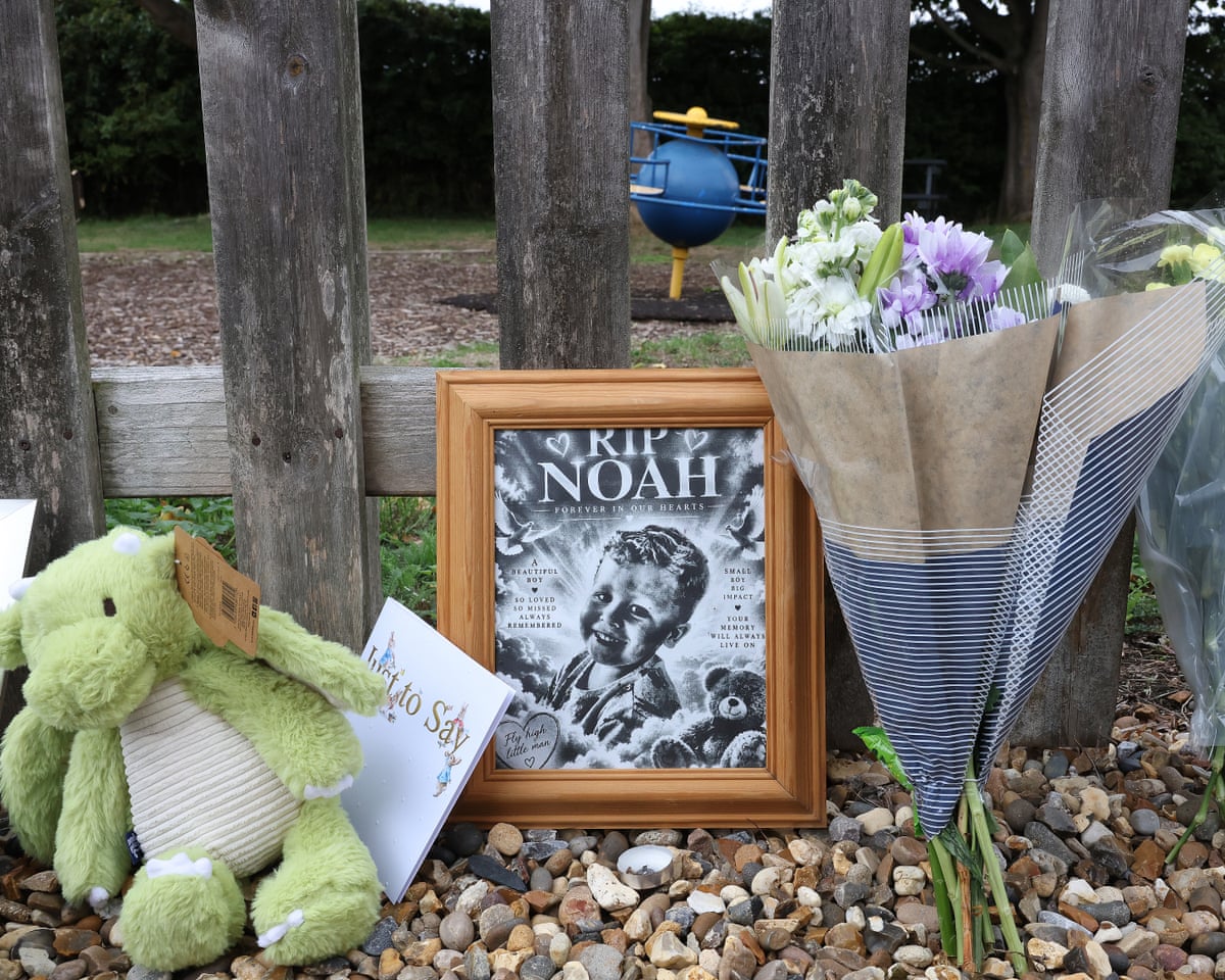 A framed AI image of Noah with the words ‘RIP Noah’ is propped against the fence, next to a bunch of flowers and a green teddy