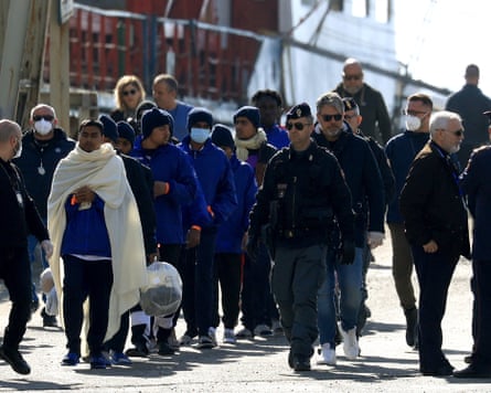 People dressed in blue jackets with blankets around their shoulders are escorted off a boat by Italian authorities
