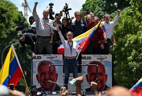 A woman waves and holds the Venezuelan flag from the center of a truck filled with people and decorated with political posters