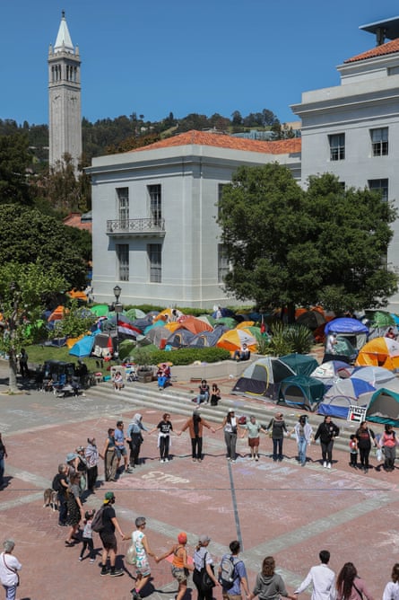 Community members gather in a circle on the berkeley campus, with tents behind them and a tower at the top left