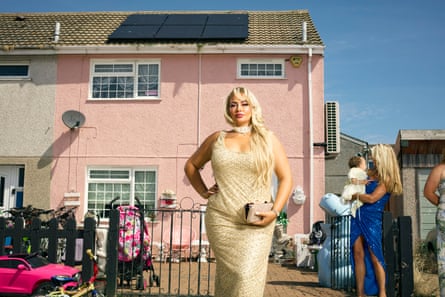 A glamorously dressed woman stands outside a pastel-pink terraced house