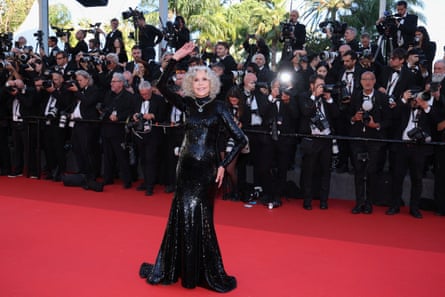 Jane Fonda waves to the cameras on the red carpet. She wears a long-sleeved, clinging floor-length black dress covered in sequins, which flares out from the knees.