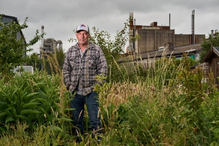 Hodson, wearing a cap, stands on an allotment with industrial buildings in the background