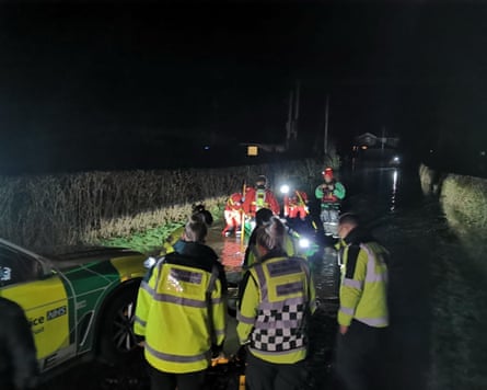 Rescue workers in hi-vis jackets shining torches on a flooded road between hedges leading to a house