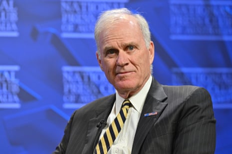 A man white hair and a dark suit jacket before a blue background.