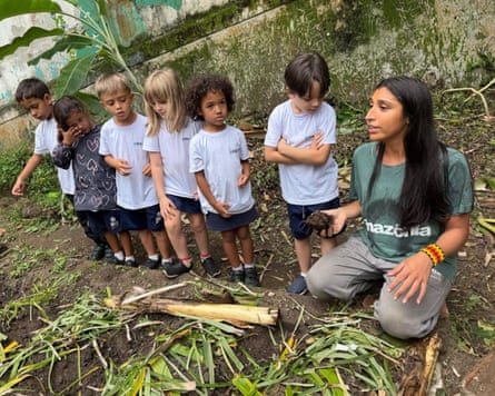A woman in a green t-shirt kneels on the ground by rows of plants, with a line of children next to her.