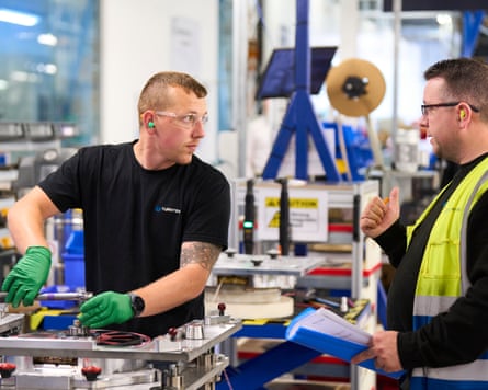 Two factory workers, one on left in protective glasses and a black T-shirt, the other on right in a hi-vis waistcoat, in discussion on the production line.