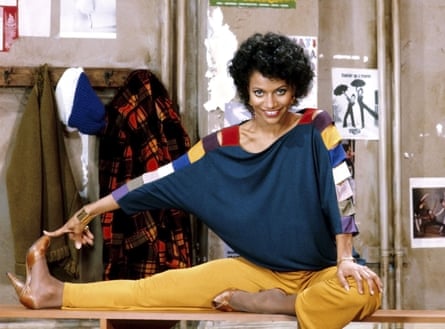 Sitting on a low bench, with one leg fully extended and the other tucked into her groin, she smiles at the camera