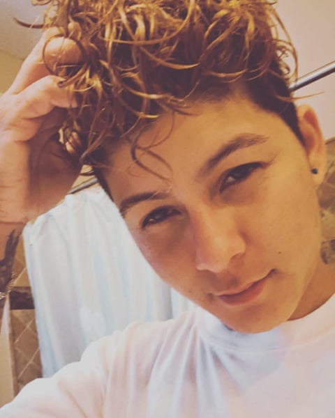 A selfie of a person with one hand in wavy blond-tipped hair, wearing a white T-shirt.