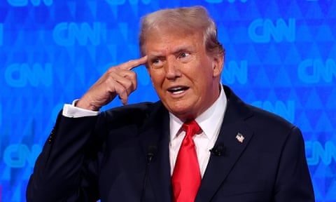 A man wearing a navy suit and red tie, Donald Trump, points to his head as he speaks