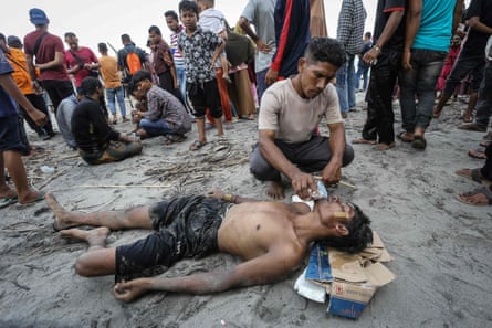 A man lies on the wet sand while another man gives him water