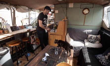 Man in the living room / kitchen of his houseboat. A cat walks across the coffee table.