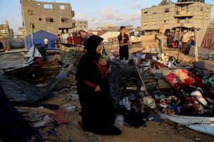 A woman holding a hand up to her heart as she looks at a pile of furniture and belongings in the rubble