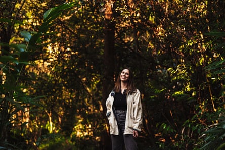 A woman stands in a clearing in the forest with a backdrop of golden and green light through trees