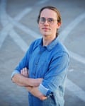 Man in denim shirt and round glasses with arms folded looks up at the camera